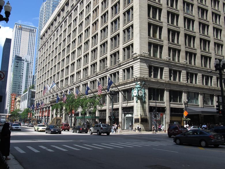 image from across the street of a large tall tan building with a green exterior clock hanging from its corner