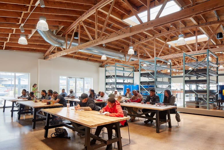 Bright room with long tables and people seated at the all facing the leftside of the frame in a room with tall ceilings and wooden beams