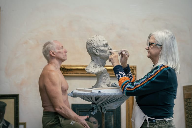 Image of a man posing looking forward to the left of the frame while a woman with white hair and glasses faces forward to the left of the frame sculpting his bust our of gray clay. 