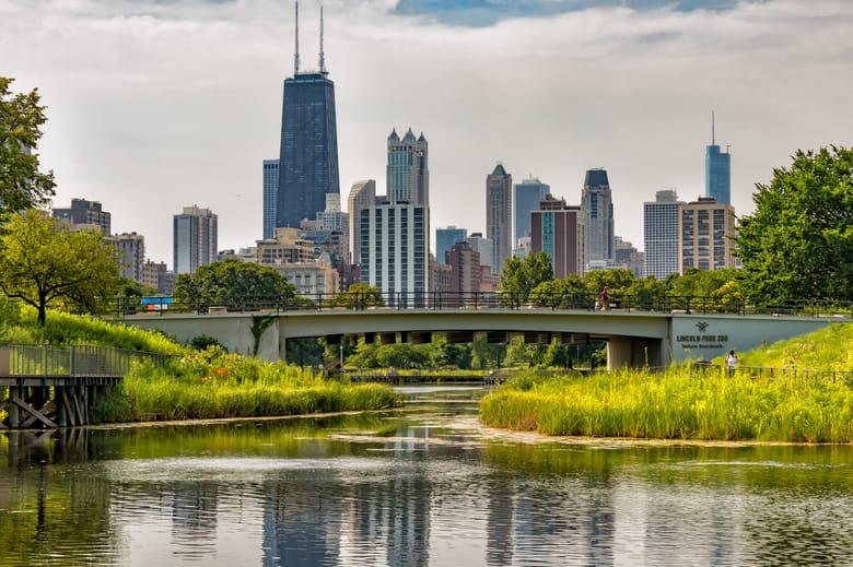 Image of a city skyline with various buildings of different heights in the background and a bridge over a river bordered by green space in the foreground