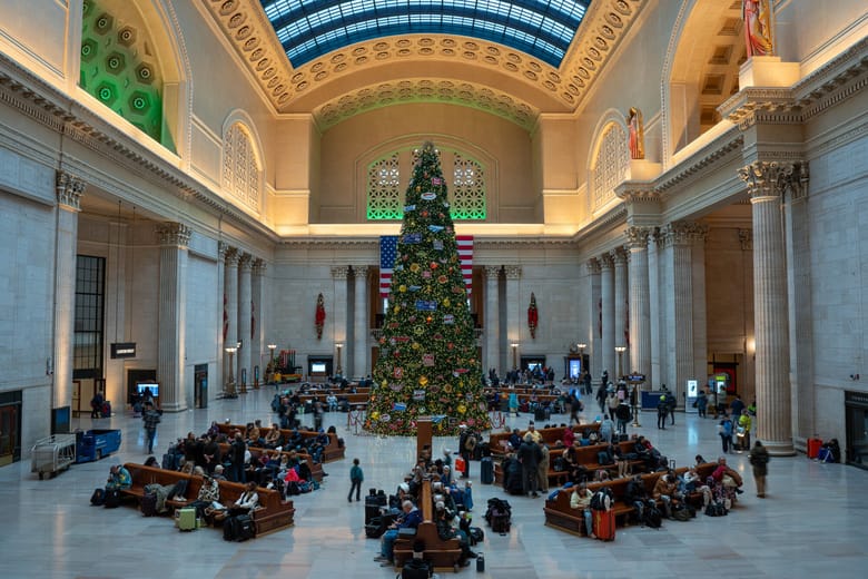 Image of a large room with high arched ceilings lit up and a large Christmas tree in the middle surrounded by people sitting on benches.