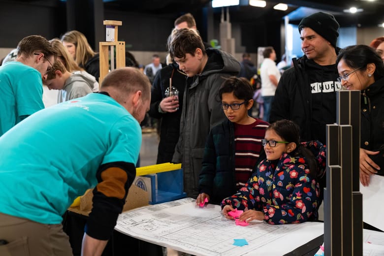 Image of two children looking over blueprints with a man in a teal shirt standing across the table from them. 