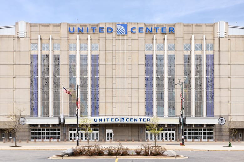 Front façade of the United Center in Chicago, viewed from across the street. The arena’s pale stone exterior features tall vertical window panels and prominent blue “UNITED CENTER” signage above the main entrance.
