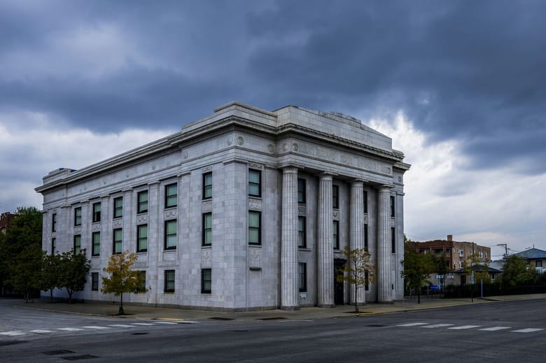 Large 3 story grey building with columns at the front and a dark cloud in the sky behind it. 