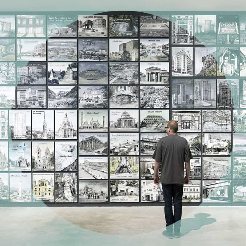 Man standing and facing away from the viewer looking at a wall of illustrations of different locations.
